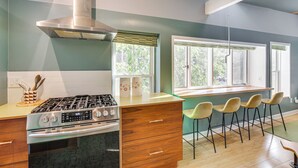 A modern kitchen with a gas stove, oven, and stainless steel range hood. There are bar stools and a window with blinds. - 3 Mi to Boothbay Harbor: Coastal Colorful Home! (East Boothbay)