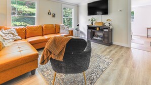 A living room with a leather sofa, a dark armchair, a fireplace, and a flat-screen TV mounted on the wall. - Peaceful Brookside Getaway ~10 Mi to Okemo Slopes! (Chester)