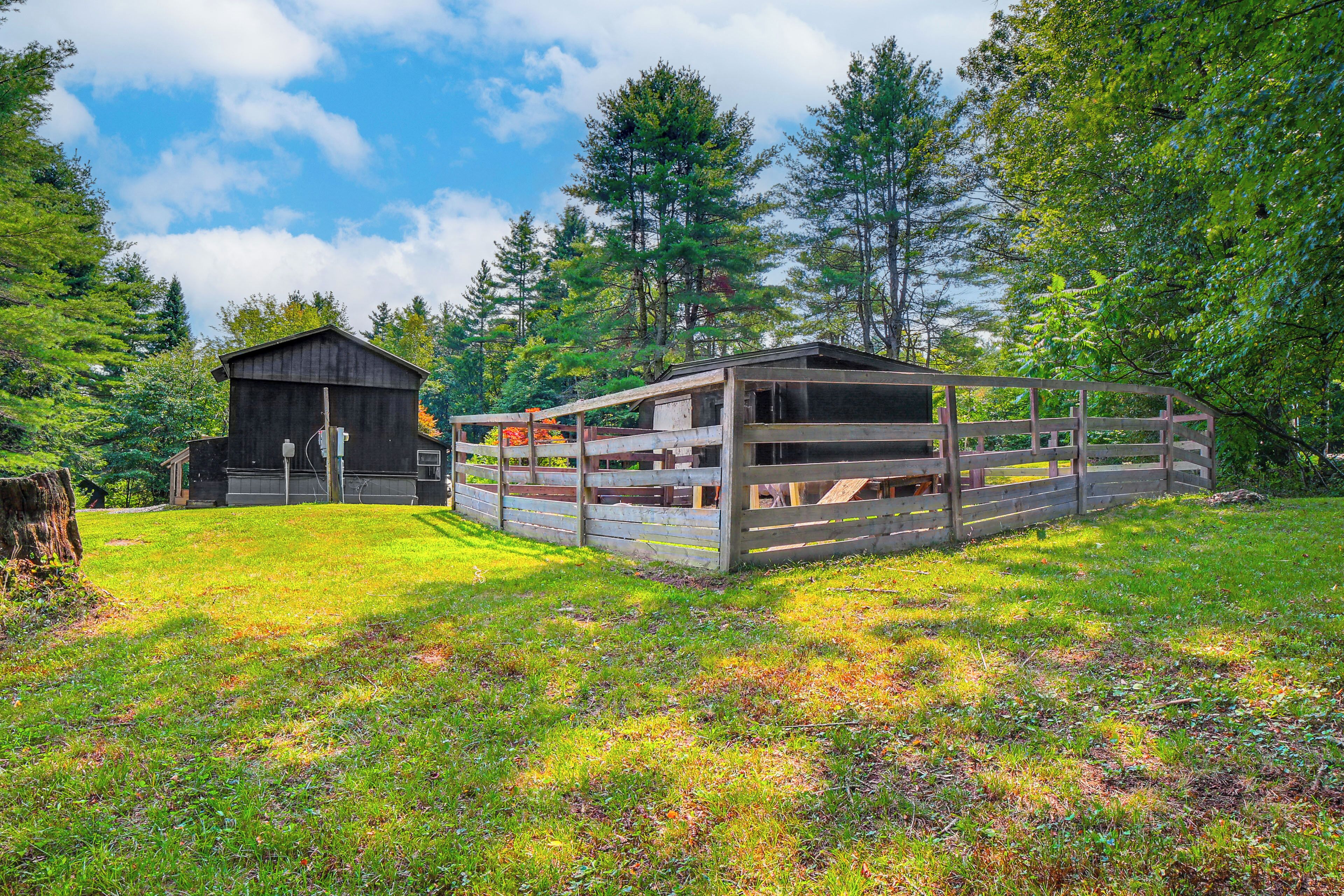 A rustic cabin with a wooden deck and a fenced area, surrounded by lush greenery and tall trees.