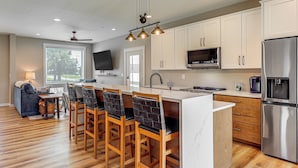 A modern kitchen with wooden bar stools, a large island, stainless steel appliances, and a flat-screen TV. - Rock Hall Retreat w/ Balcony Near Park & Marina! (Rock Hall)