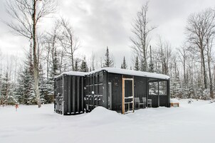 Una cabaña negra en un bosque nevado con una puerta y un banco afuera.