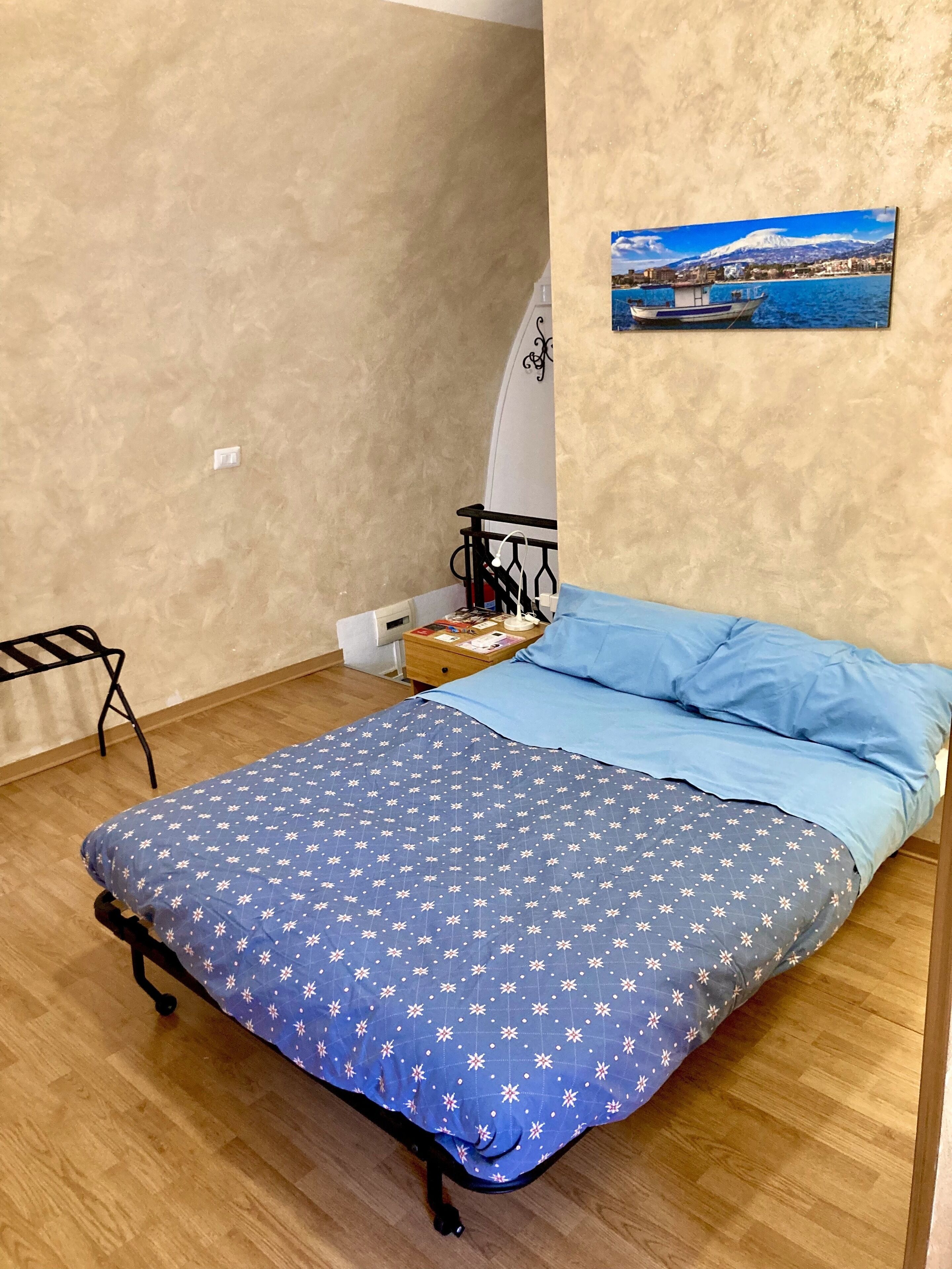 A bedroom with a bed, a small table with a lamp, a chair, and a picture on the wall.