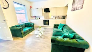 A living room with two green sofas, a coffee table, and a kitchen area with cabinets and appliances. - 5bed-10 Guests-near City Centre-free Parking (Leeds)