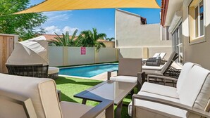 A poolside area with lounge chairs and a table, a covered grill, and a yellow shade sail. - Aruba Oasis! 3BR Home w Pool & BBQ near Palm Beach (Noord)