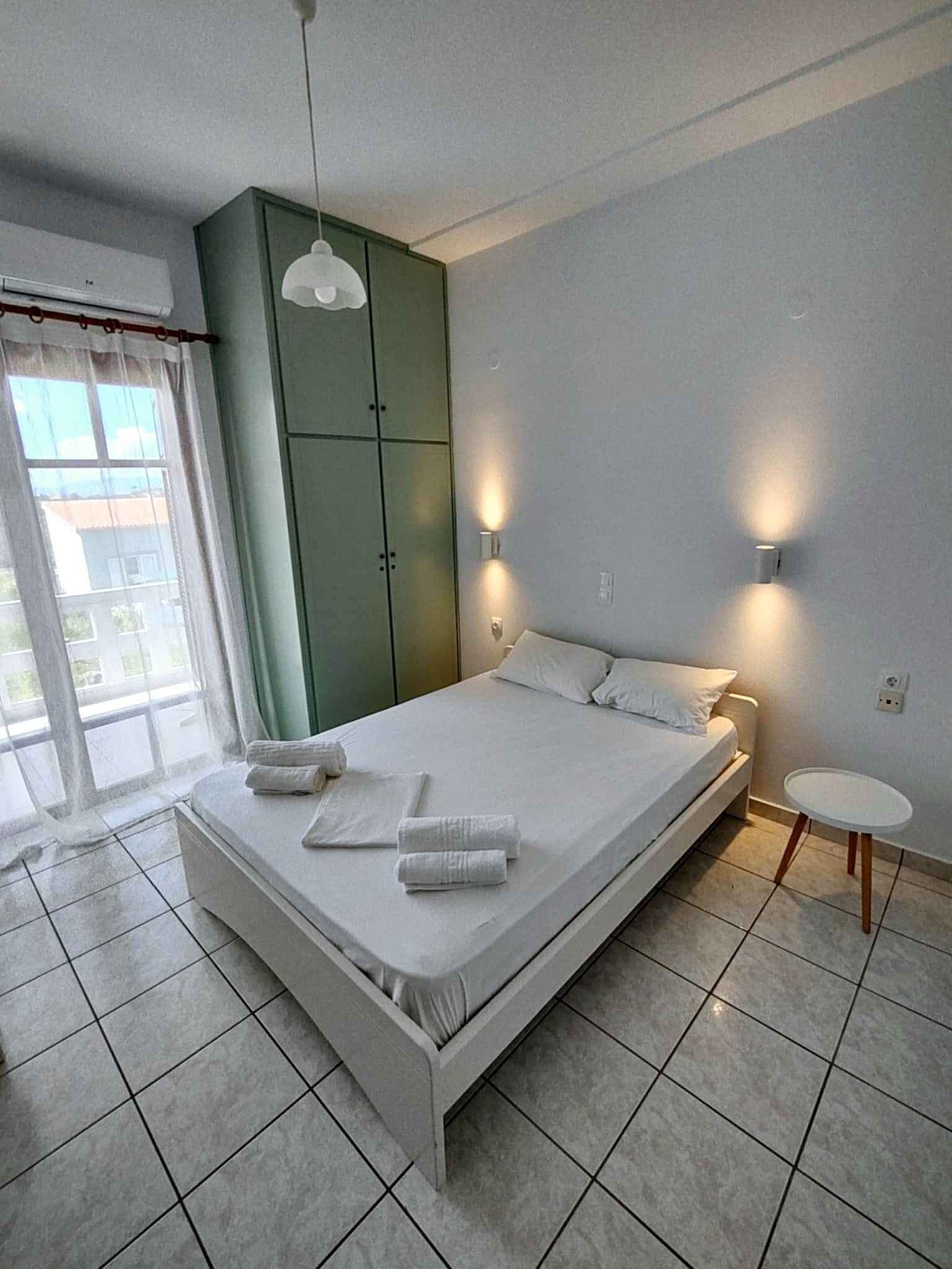 A modern bedroom with a bed, a green wardrobe, a small table, and a window with curtains.
