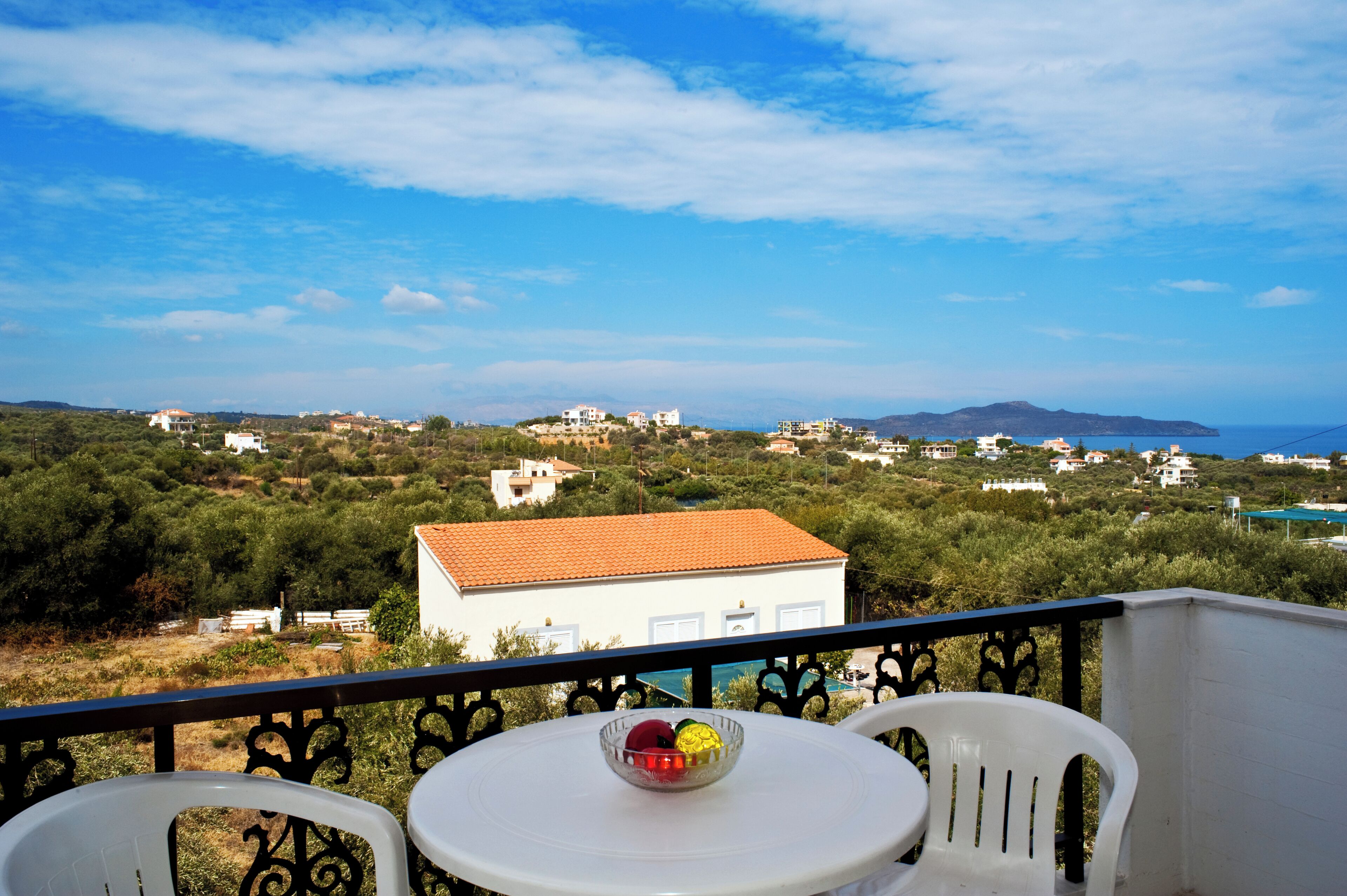 Un balcón con una mesa y sillas, un frutero y vistas a un pueblo costero con edificios y zonas verdes.