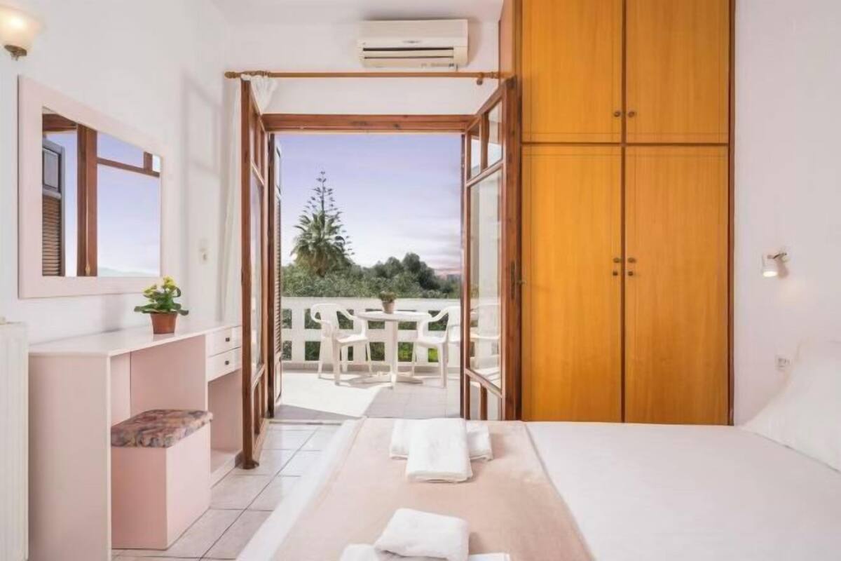 A bedroom with a bed, a wooden wardrobe, a small desk with a chair, and a view of the outdoors.