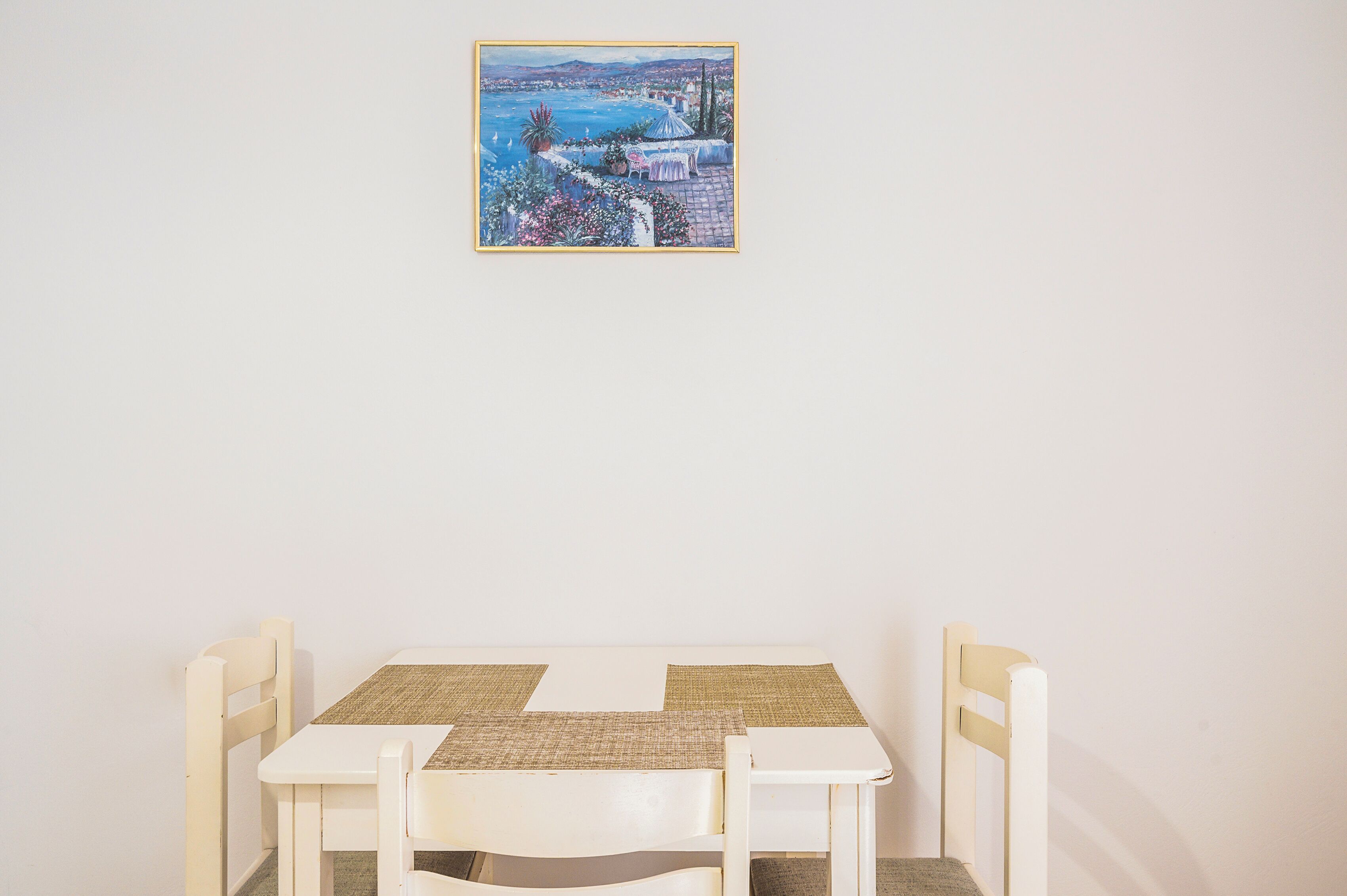 Een witte tafel met twee stoelen en een geweven placemat, daarboven een lijst met een schilderij van een kustlandschap, en een effen witte muur.