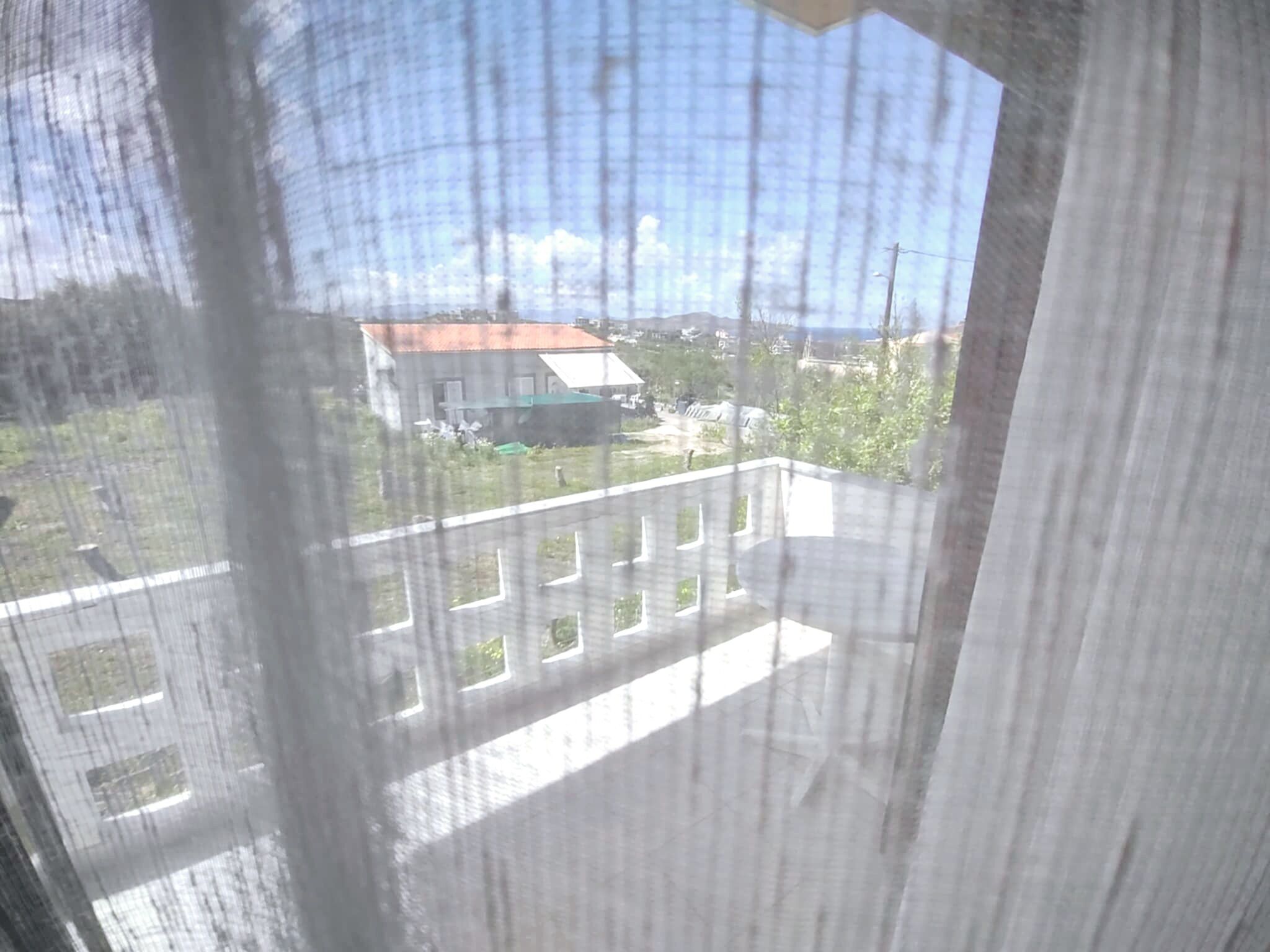 A view through a window with a white curtain, featuring a building with a red roof and a green lawn.