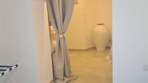 Un couloir étroit, orné d’un rideau beige noué en haut, un vase blanc posé sur le sol et une chaise blanche avec un coussin à rayures.