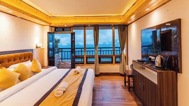 A hotel room with a large bed, wooden ceiling, a balcony with a view, a TV, and a kitchenette. - The Golden Retreat Hotel in Pelling (Gyalshing)