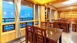 A dining area with wooden tables and chairs, large windows with curtains, and a view of a body of water. - The Golden Retreat Hotel in Pelling (Gyalshing)