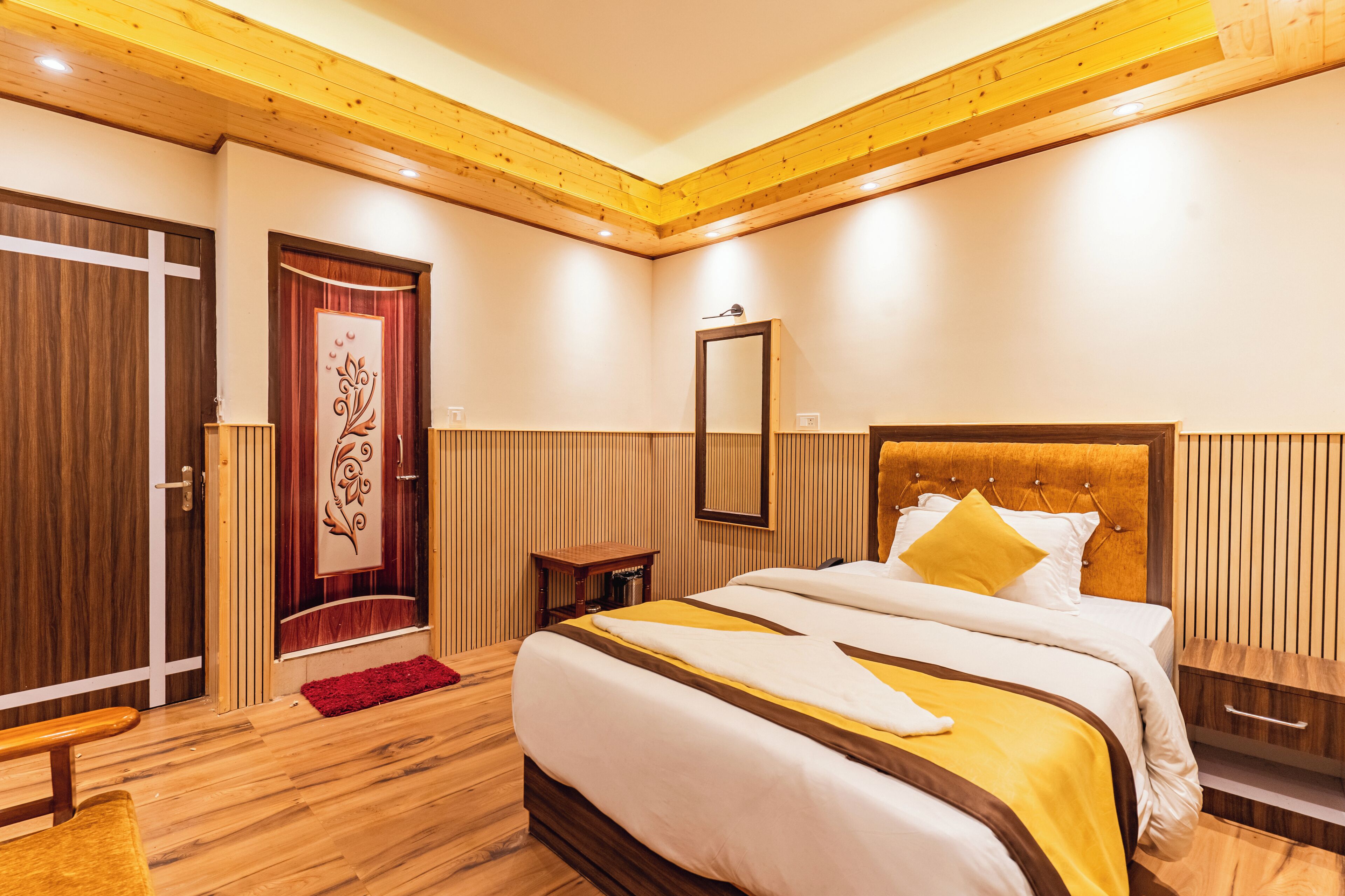 A hotel room with a wooden ceiling, a large bed with a yellow and white bedspread, a wooden nightstand, a bedside table, and a wooden wardrobe with a cross design.