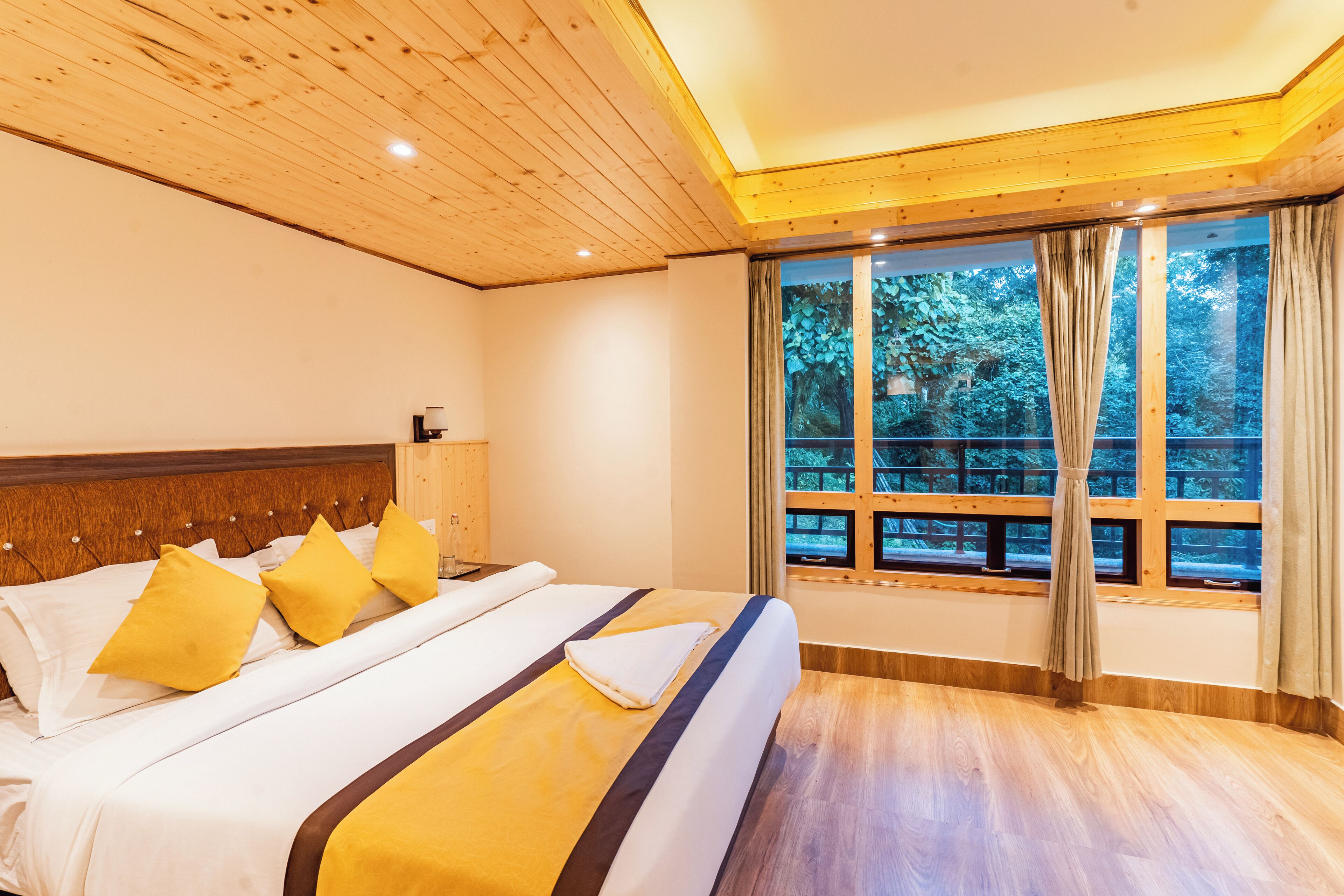 A bedroom with a wooden ceiling, a large window with curtains, a bed with white and yellow bedding, and a wooden floor.