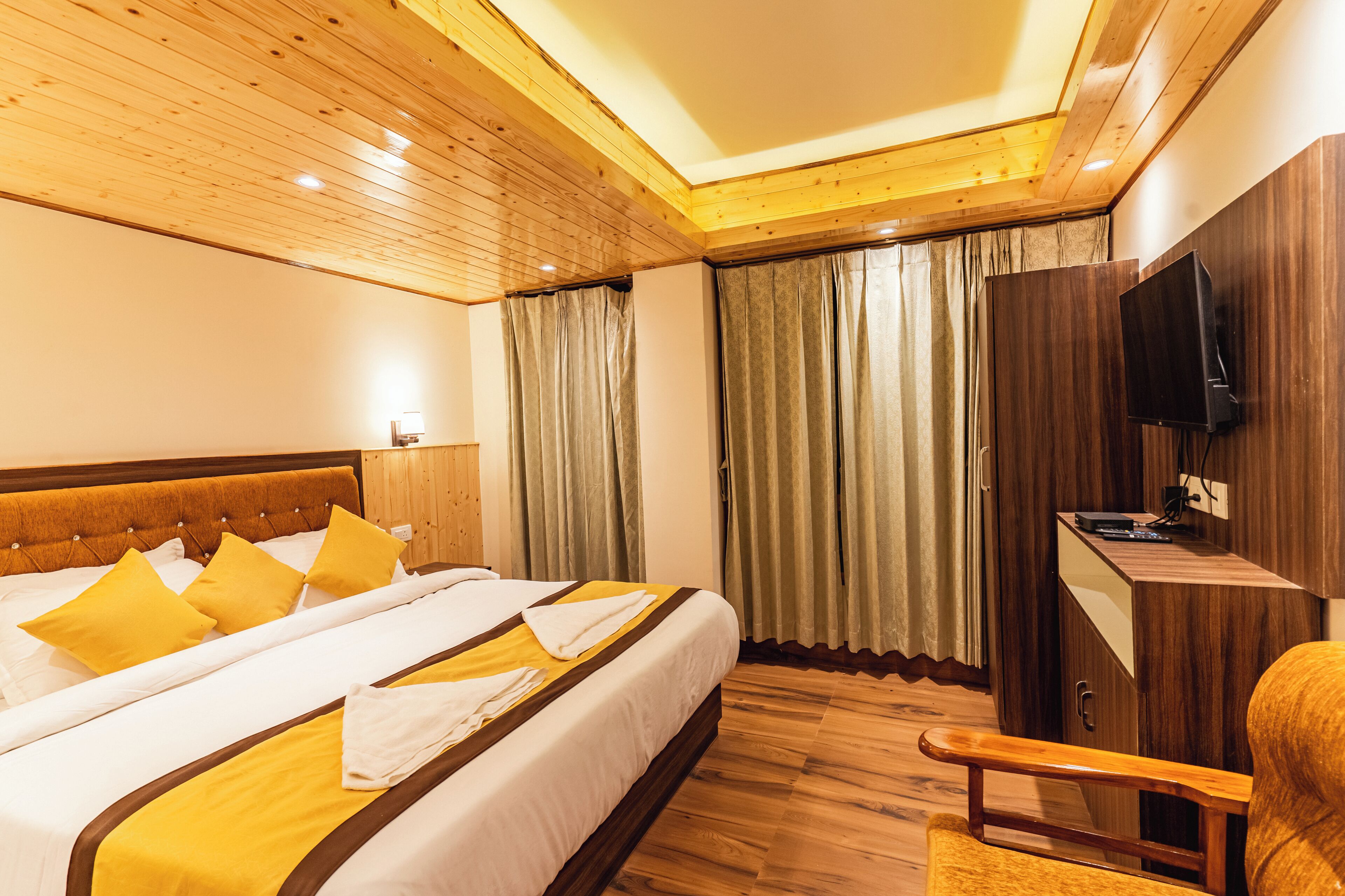 A hotel room with a wooden ceiling, a large bed with yellow and white bedding, a desk with a flat-screen TV, and a wooden chair.