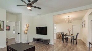 Un salón amplio con una mesa de centro de madera oscura, un televisor de pantalla plana montado en la pared y una zona de comedor con una araña.