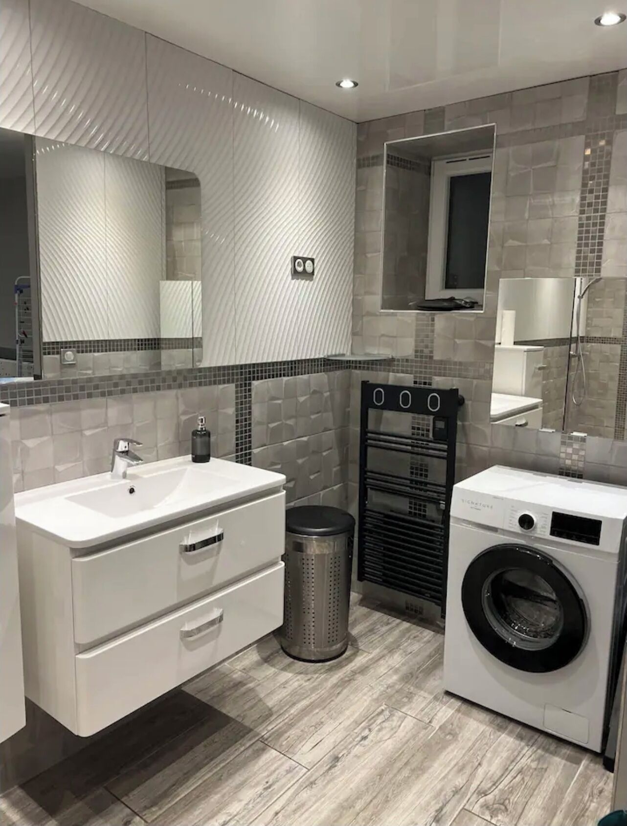 Une salle de bain moderne avec un grand miroir, un lavabo, un lave-linge et une poubelle.
