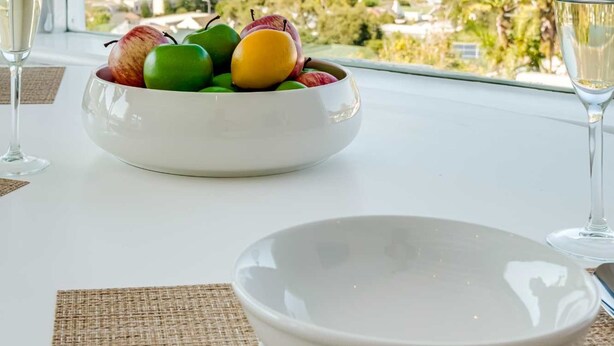 Una mesa de comedor con un bol de fruta y dos vasos de agua, con vistas a un paisaje pintoresco de montañas y una masa de agua.