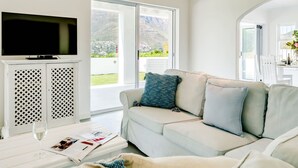 Una sala de estar con un televisor de pantalla plana sobre un mueble blanco, un sofá beige con cojines, y una mesa con una copa de vino y revistas.