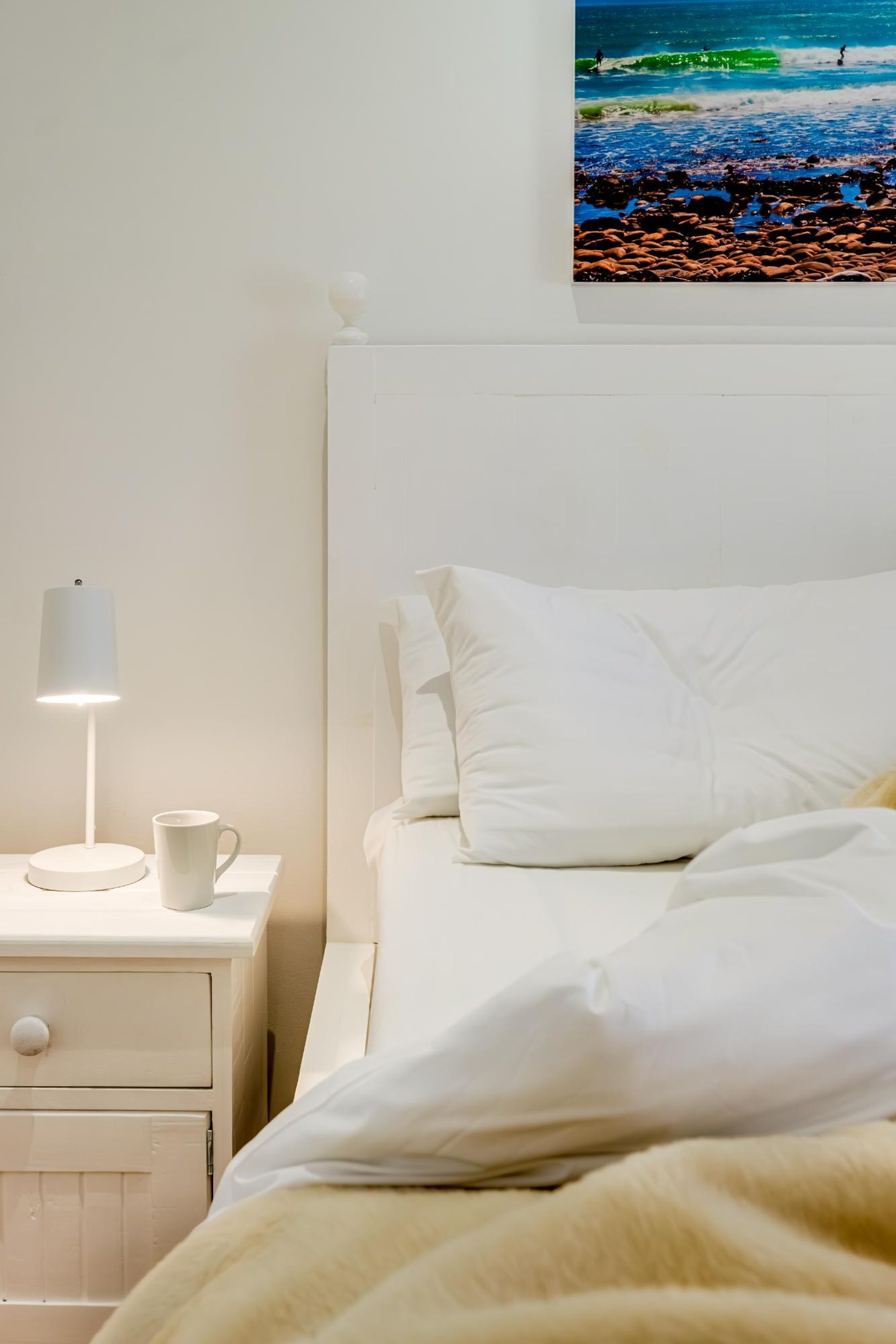 A neatly made bed with white linens and a beige blanket, a bedside table with a lamp and a mug, and a wall-mounted picture of a beach scene.