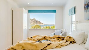 Un dormitorio con cama, ventana con vistas a la montaña y al mar, y cabecera blanca.