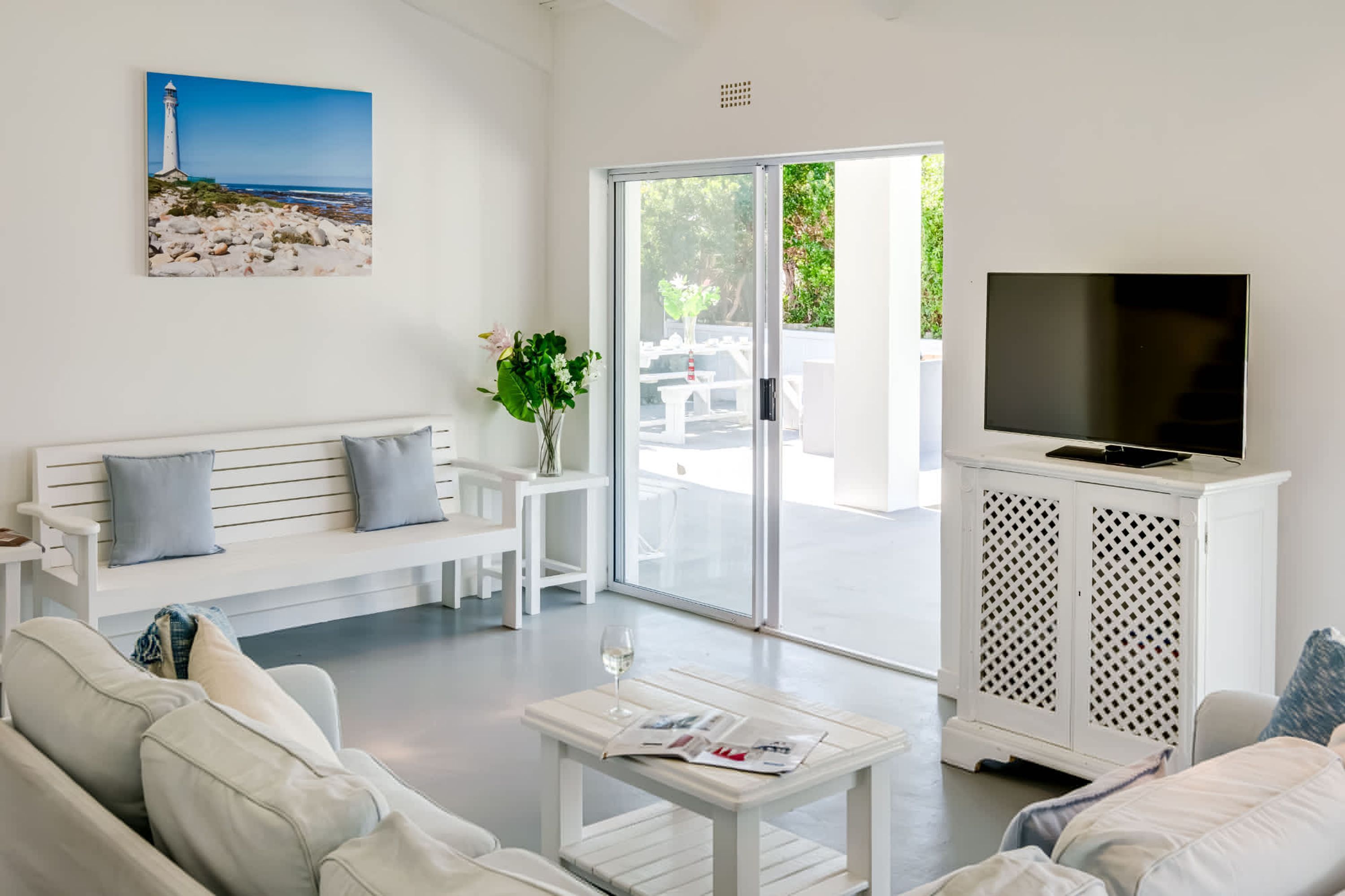 A modern living room with a white sofa, a coffee table with magazines, a TV on a white cabinet, and a painting of a lighthouse on the wall.