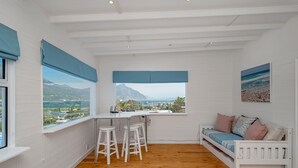 Una habitación con piso de madera, paredes blancas, un sofá blanco, una mesa de comedor con taburetes y ventanas amplias con cortinas azules.