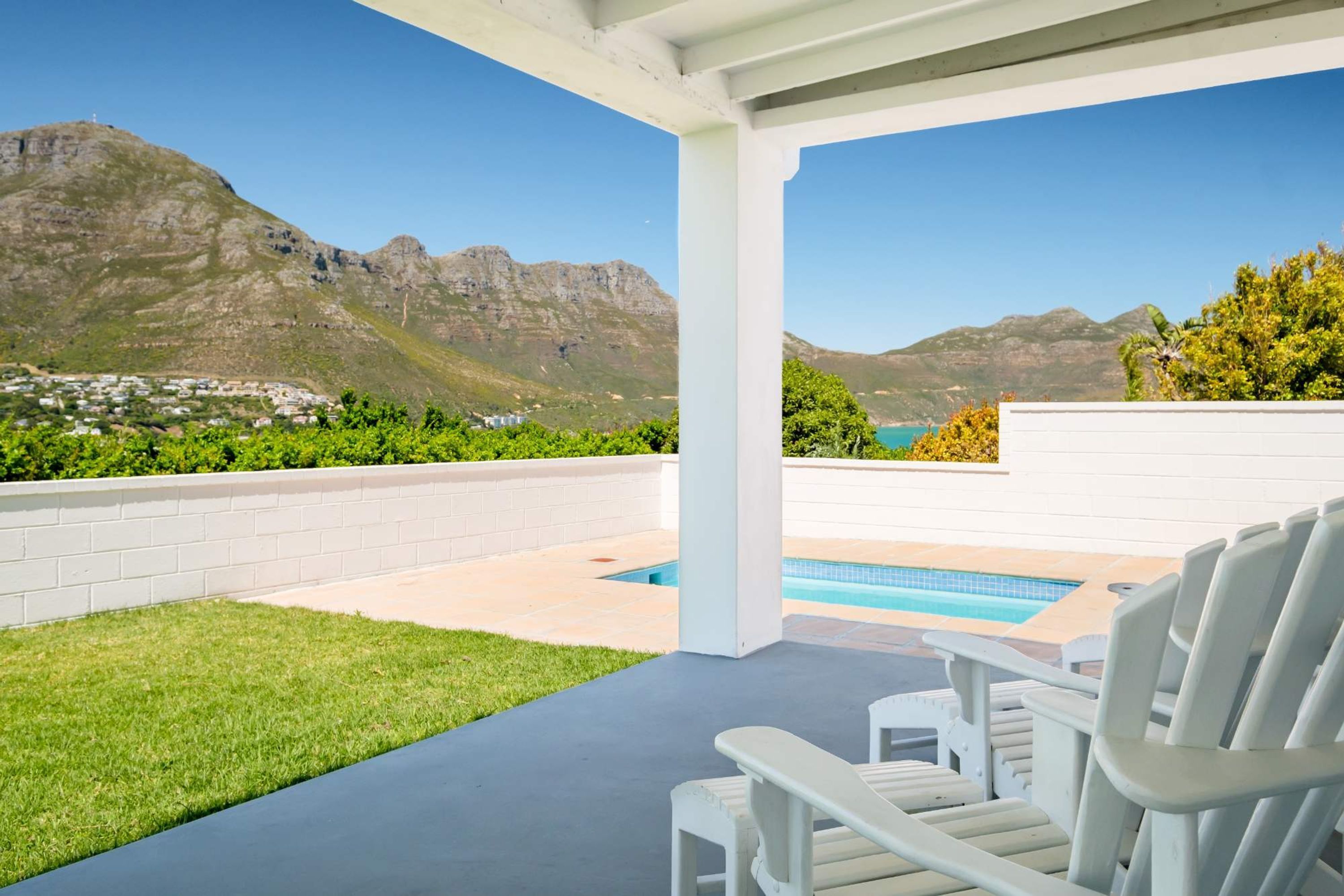 Un espacio exterior techado con piscina, sillas blancas y vistas a montañas y un pueblo.