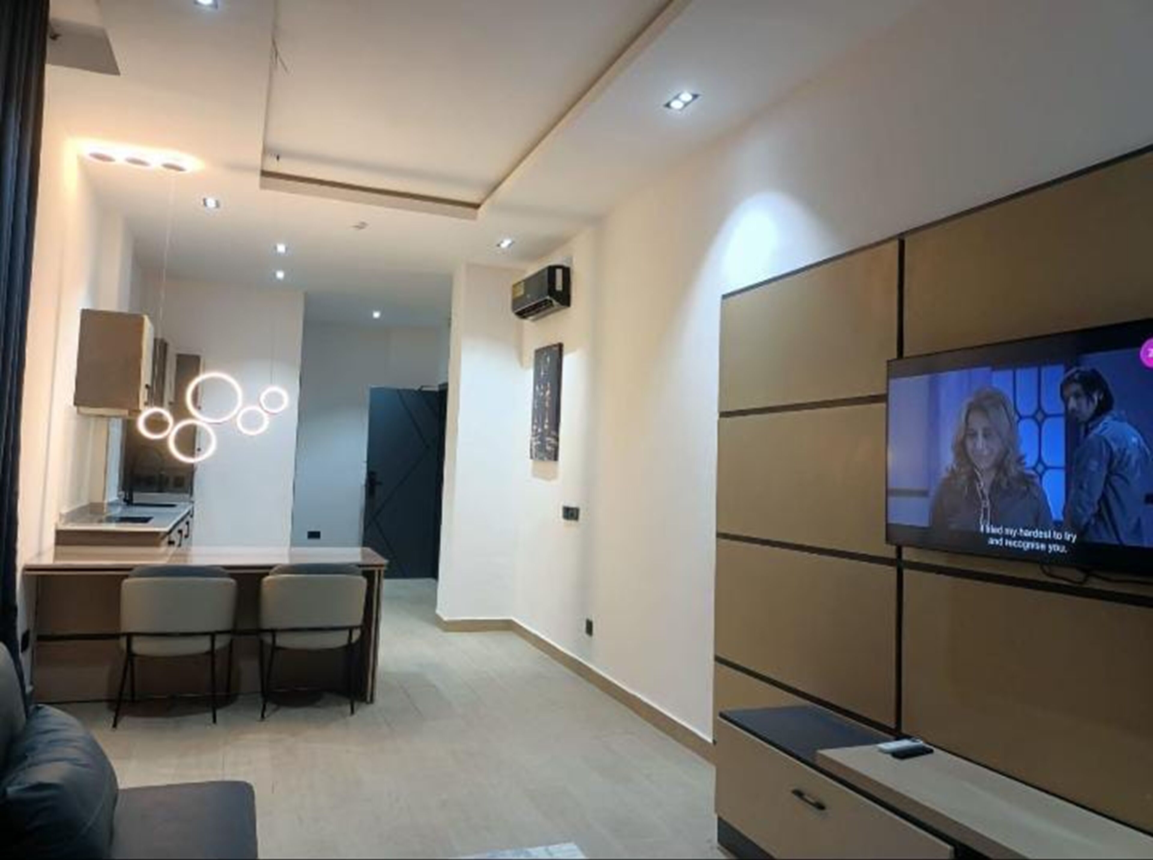 A modern living room with a flat-screen TV mounted on the wall, a dining area with a small table and chairs, and a kitchen area with cabinets and a countertop.