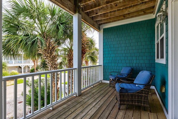 A wooden deck with two wicker chairs, a teal brick wall, and palm trees in the background.