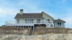 A beachfront house with a large deck, multiple windows, and a staircase leading to a second level. - Dune Crest - Southern Shores, Oceanfront, Pool, Hot Tub, Pool Table, Pets OK (Southern Shores)