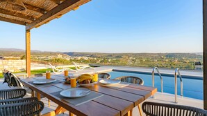 A wooden table with plates, glasses, and a fruit bowl set for breakfast. A pool with a view of the countryside is visible in the background.