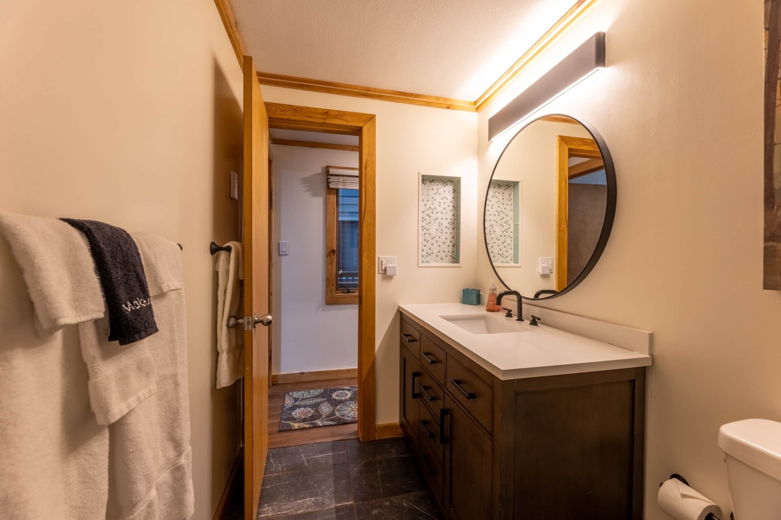 A bathroom with a wooden vanity, a round mirror, a toilet, and a towel rack.