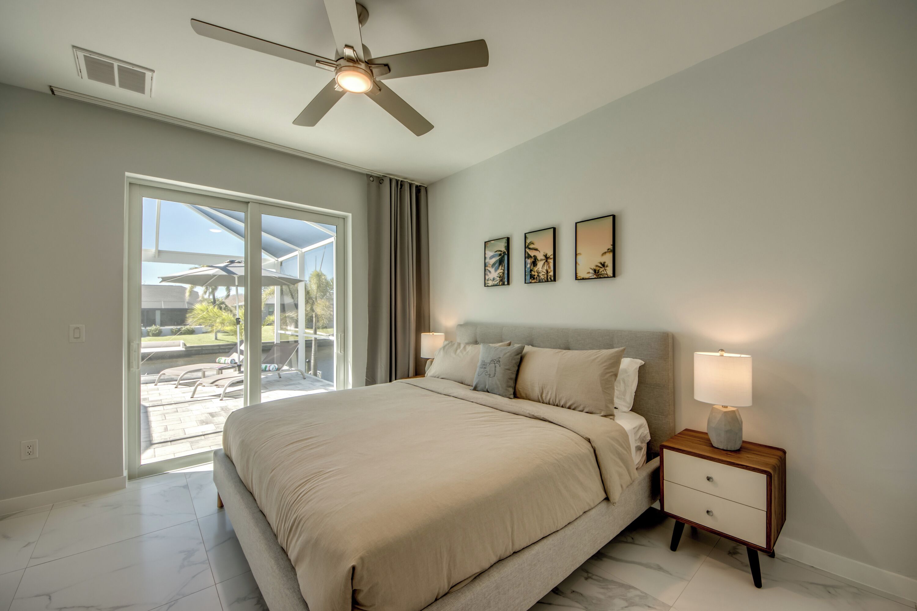 Quarto com cama, mesa de cabeceira com abajur, ventilador de teto e vista para o exterior.