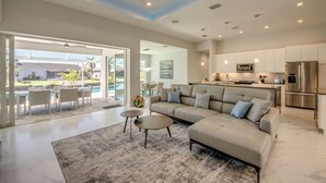A spacious living area with a modern sofa, a coffee table, and a dining set by a large sliding glass door leading to a patio. - Villa Summer Feeling - newly built, on water, pool & spa (Cape Coral)