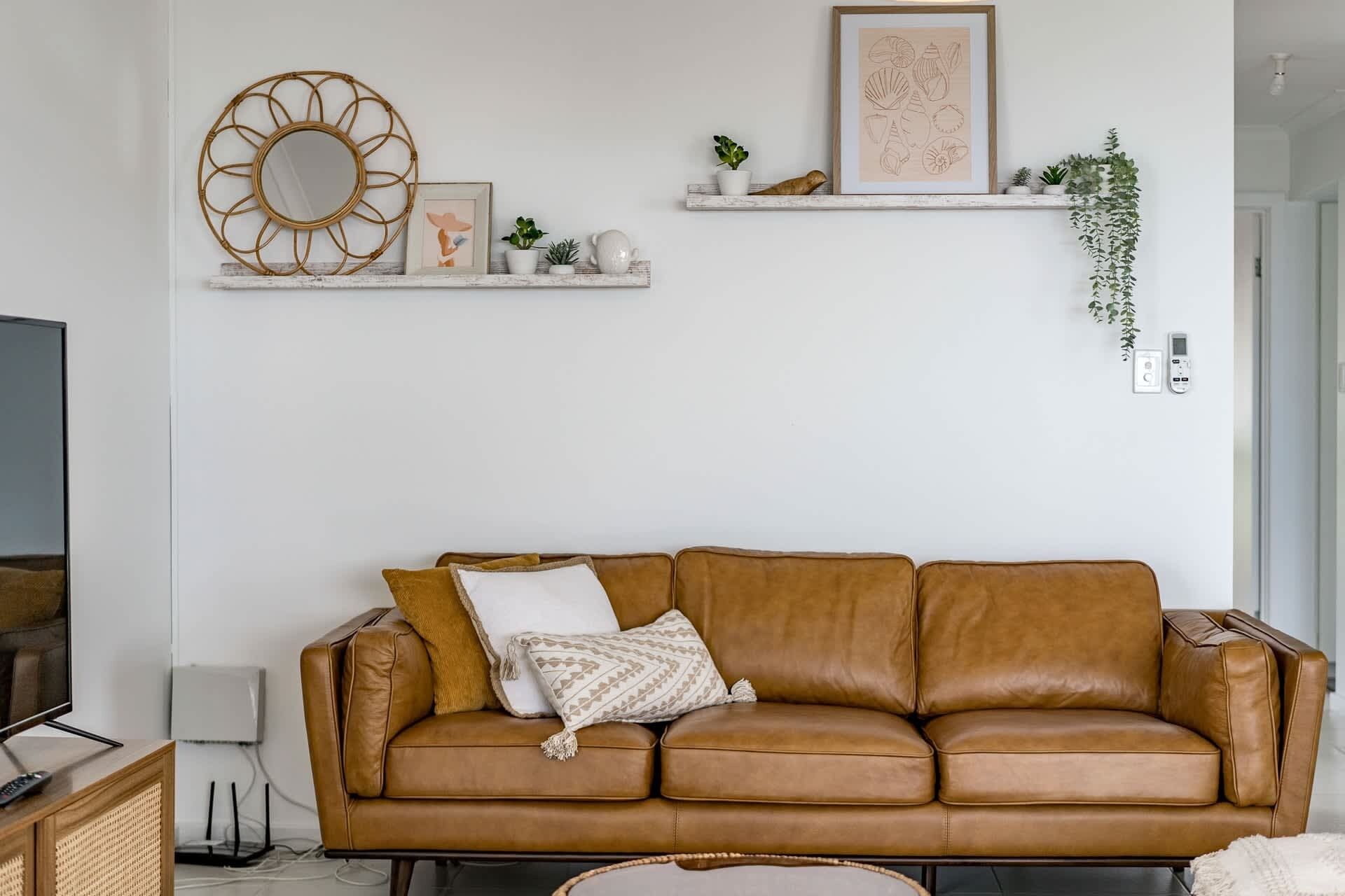Een bruine leren bank met kussens, een wandspiegel, een lijst met een afbeelding, en een plank met planten en decoratieve objecten.