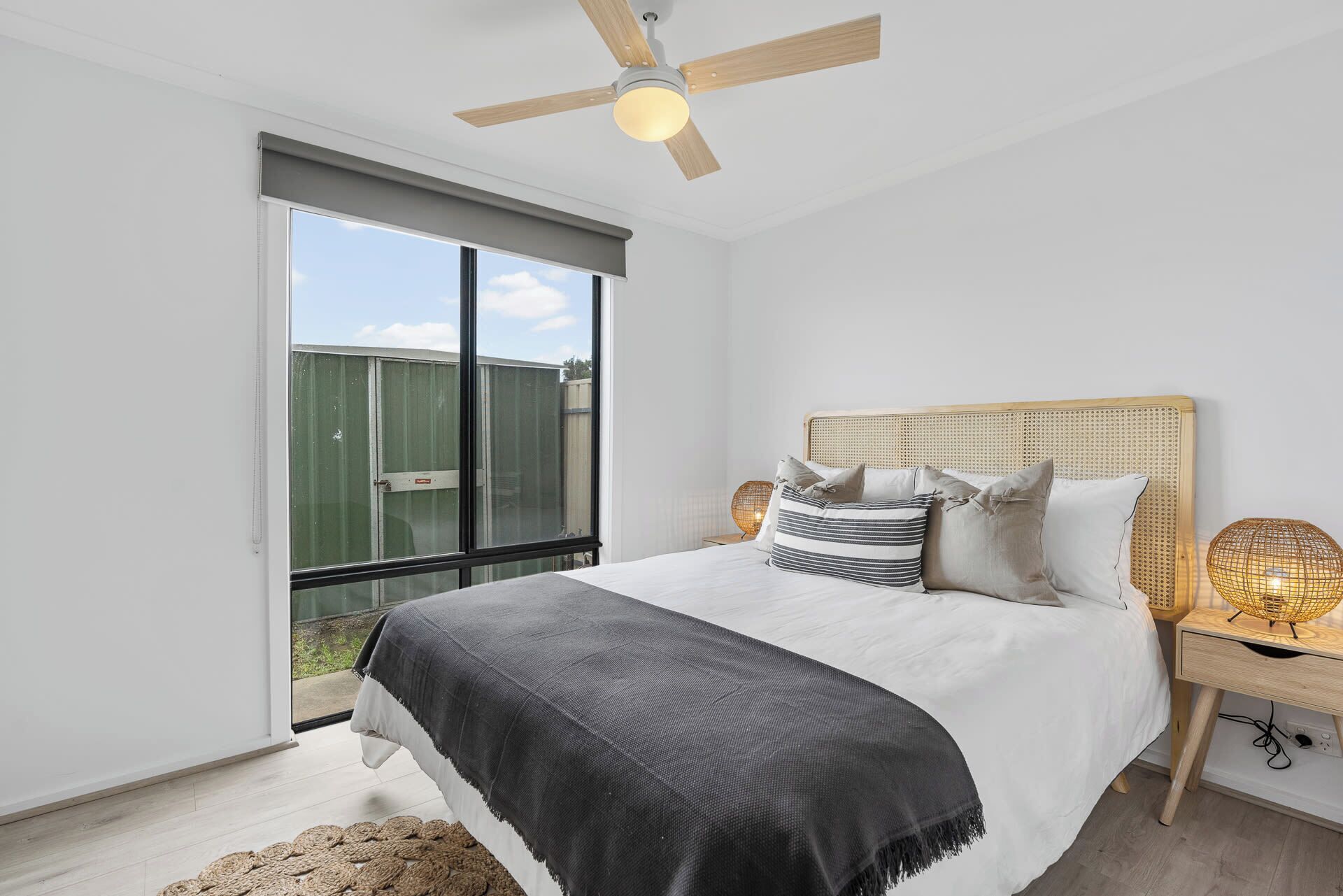A bedroom with a bed, a ceiling fan, a bedside table with a lamp, and a view of the outside.