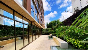 A modern building with large glass windows, a wooden deck, and a garden area with plants and a water feature. - Flat FM20 Vista Lateral - By Almare (João Pessoa)