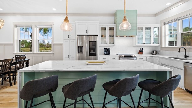 Uma cozinha moderna com armários brancos, uma ilha grande com banquetas, eletrodomésticos de aço inoxidável e iluminação pendente.
