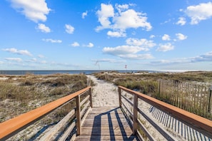 A wooden boardwalk leading to a beach with sand and sparse vegetation, under a clear sky.