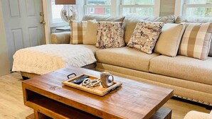 A cozy living room with a wooden coffee table, a beige sofa with patterned cushions, and a view of a window.