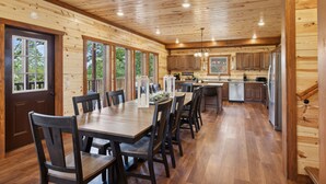 Interior de una cabaña de madera con comedor, cocina y ventanas amplias.