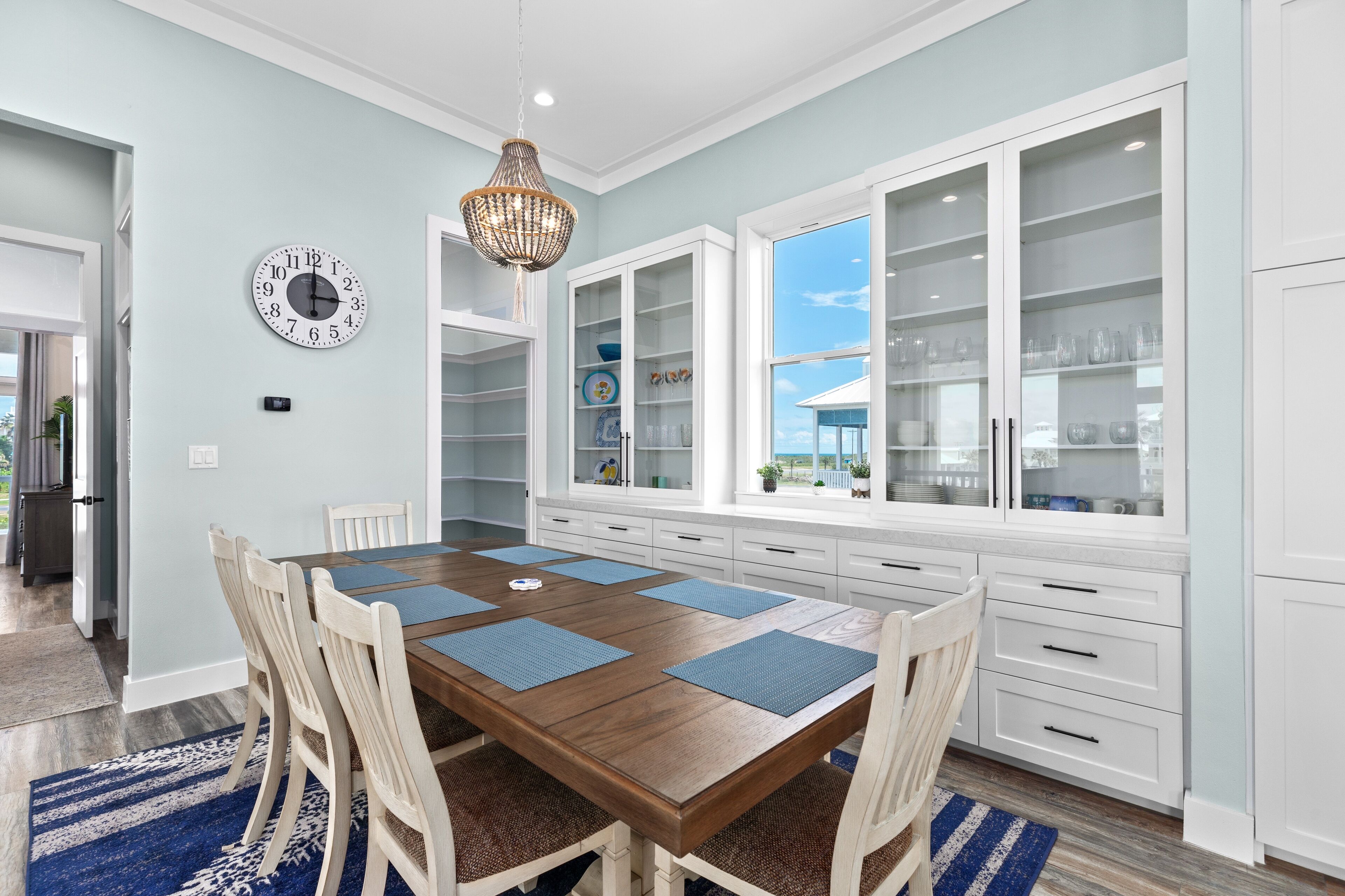 Een eetruimte met een houten tafel, blauwe placemats en witte stoelen. Er is een groot wit kastje met glazen deuren en een wandklok.