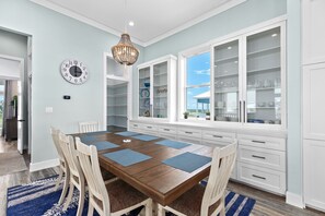 A dining area with a wooden table, blue placemats, and white chairs. There is a large white cabinet with glass doors and a wall clock. - Seaside Rendezvous - Private Home Across From Community Park 4 Bedroom Home by RedAwning (South Padre Island)