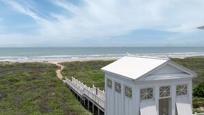 Une maison blanche située sur la plage, dotée d’une terrasse en bois qui mène à la plage et offrant une vue sur l’océan.