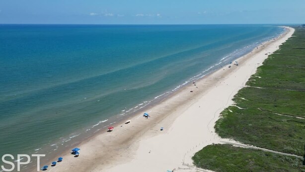 Ovenudblik over en lang, smal strand med en vej, der løber parallelt langs den, samt strandparasoller og enkelte køretøjer.
