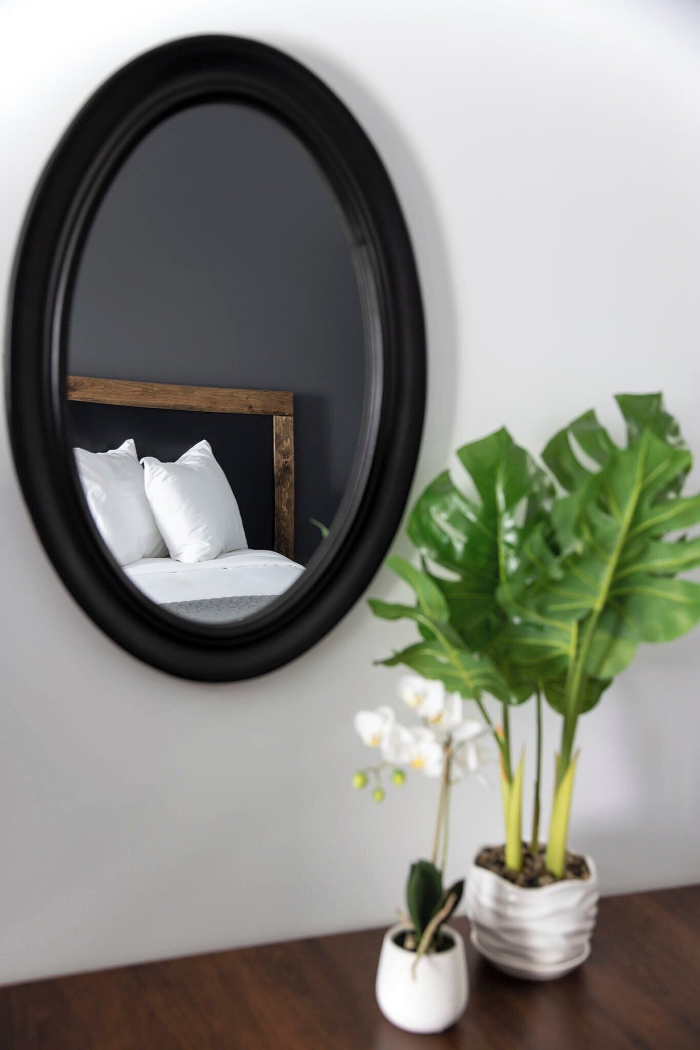 Een ronde spiegel aan een muur, die een bed met wit beddengoed en een houten hoofdeinde weerspiegelt. Er staan twee potplanten bij, één met brede bladeren en de andere met kleine witte bloemen.
