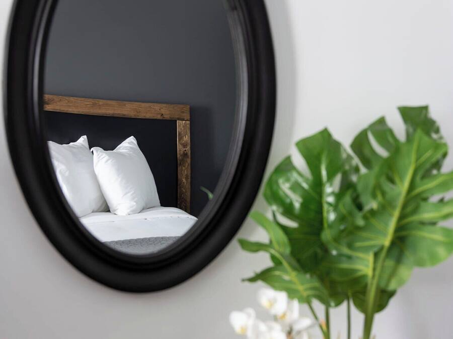 Un espejo redondo en una pared, reflejando una cama con ropa blanca y un cabecero de madera. Hay dos plantas en macetas, una con hojas grandes y otra con pequeñas flores blancas.