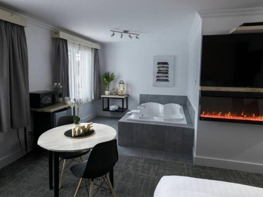 A hotel room with a bed, a TV mounted on the wall, a fireplace, a desk with a chair, and a window with curtains.