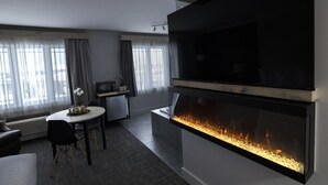 A modern living room with a large flat-screen TV, a fireplace, a dining area with a table and chairs, and a kitchen area with a microwave and a refrigerator. - Hôtel Napoléon (Gatineau)